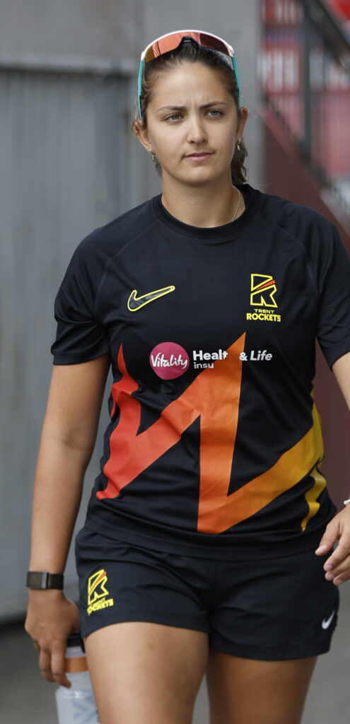 A person wearing a black sports jersey with orange and yellow accents, featuring logos for Trent Rockets and Vitality, walks while holding a water bottle.