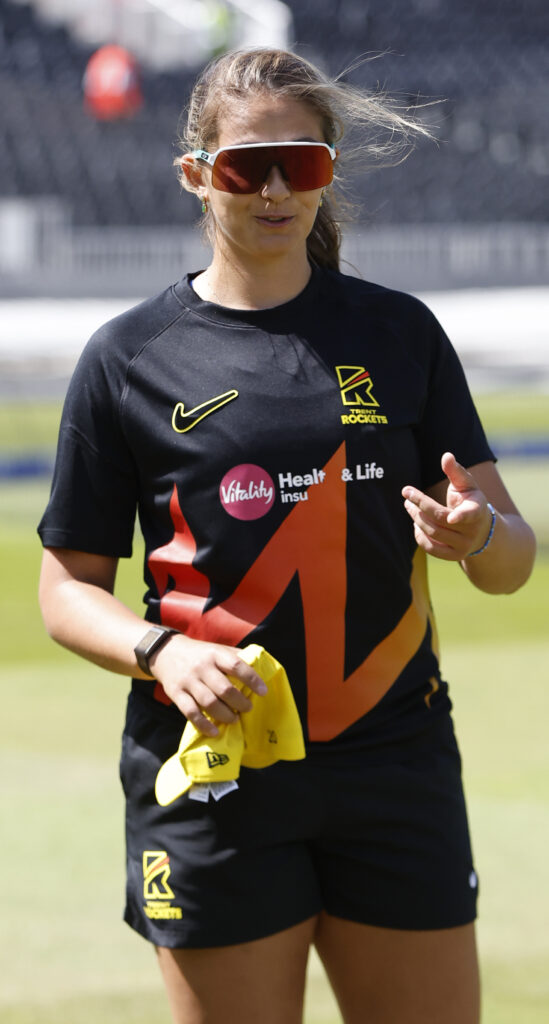 A person wearing sunglasses and a black sports jersey with orange and yellow accents walks on a grass field while holding a yellow cap.